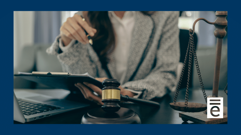 A brunette lady in a business suit sitting at a desk with a clipboard, computer, and gavel. The scales of justice are displayed to the right.